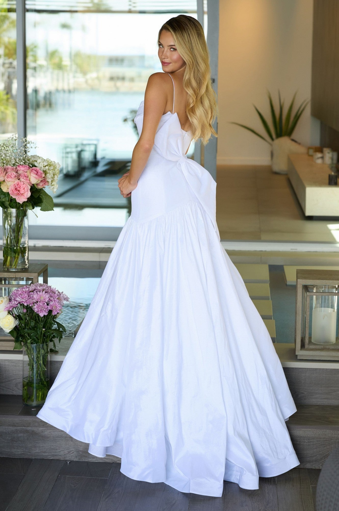 A woman wears the Ava Presley 26422 Drop-Waist Mermaid Prom Gown, standing indoors facing slightly away from the camera, with flowers and modern decor in the background.